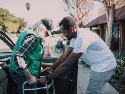 Volunteer helping an elderly man with a walking frame out of a car